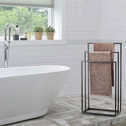 Bilder eines Handtuchständers im Badezimmer, der aus schwarzen Metallbügeln besteht und mit einem roten Gewebe überzogen ist. Ein silberner Wasserhahn ist an der Badewanne befestigt und es gibt verschiedene Artikel auf dem Fensterbrett. Eine Fensterbank mit grünen Pflanzen außerhalb sorgt für natürliches Licht, was eine entspannte Atmosphäre schafft. Der Boden ist mit Fliesen bedeckt, die ein Grau-Weiß-Muster haben. Außerdem ist im Bild 'Smartpeas Handtuchhalter Stehend' und 'Handtuchständer Schwarz Aus Rostfreiem Pulverbeschichtetem Stahl' zu lesen.